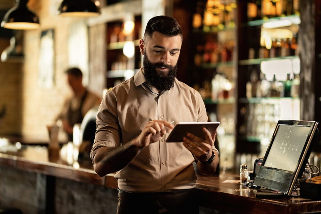 A man using a tablet, standing next to a point of sale on a counter.